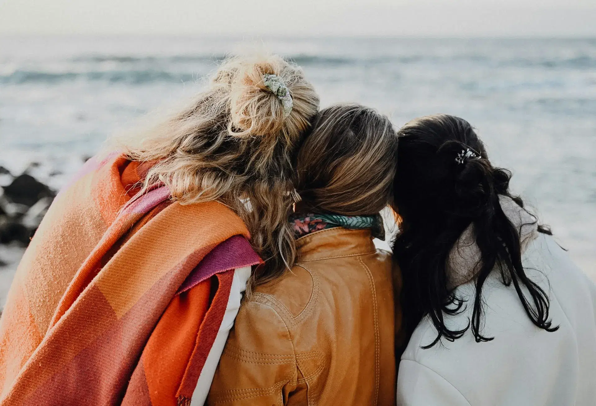 Three women sitting close together by the ocean, heads resting on one another's shoulders