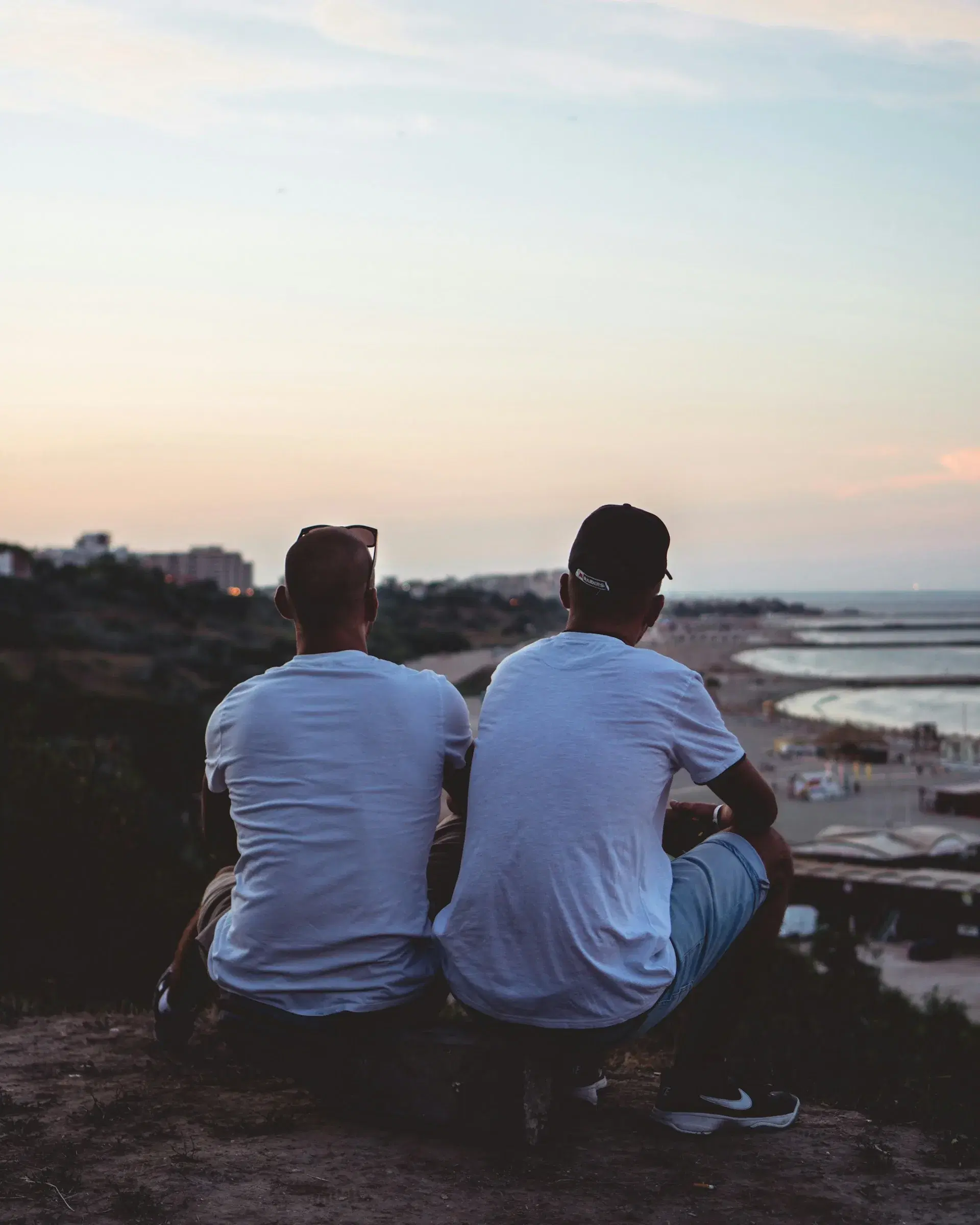 Two men sitting side by side on a hillside at dusk, looking out over the coast